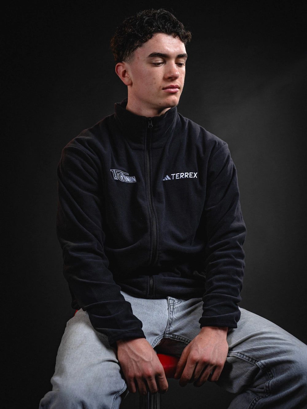 Junger Mann mit dunklem Fleece-Jacket und Jeans sitzt auf einem Stuhl vor einem dunklen Hintergrund.