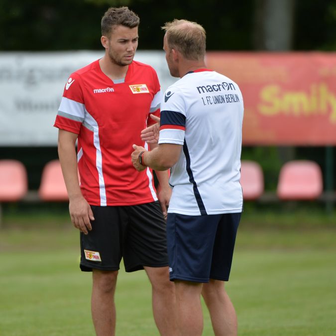 Zwei Fußballspieler im Gespräch auf einem Trainingsplatz, einer trägt ein rotes, der andere ein weißes Trikot.