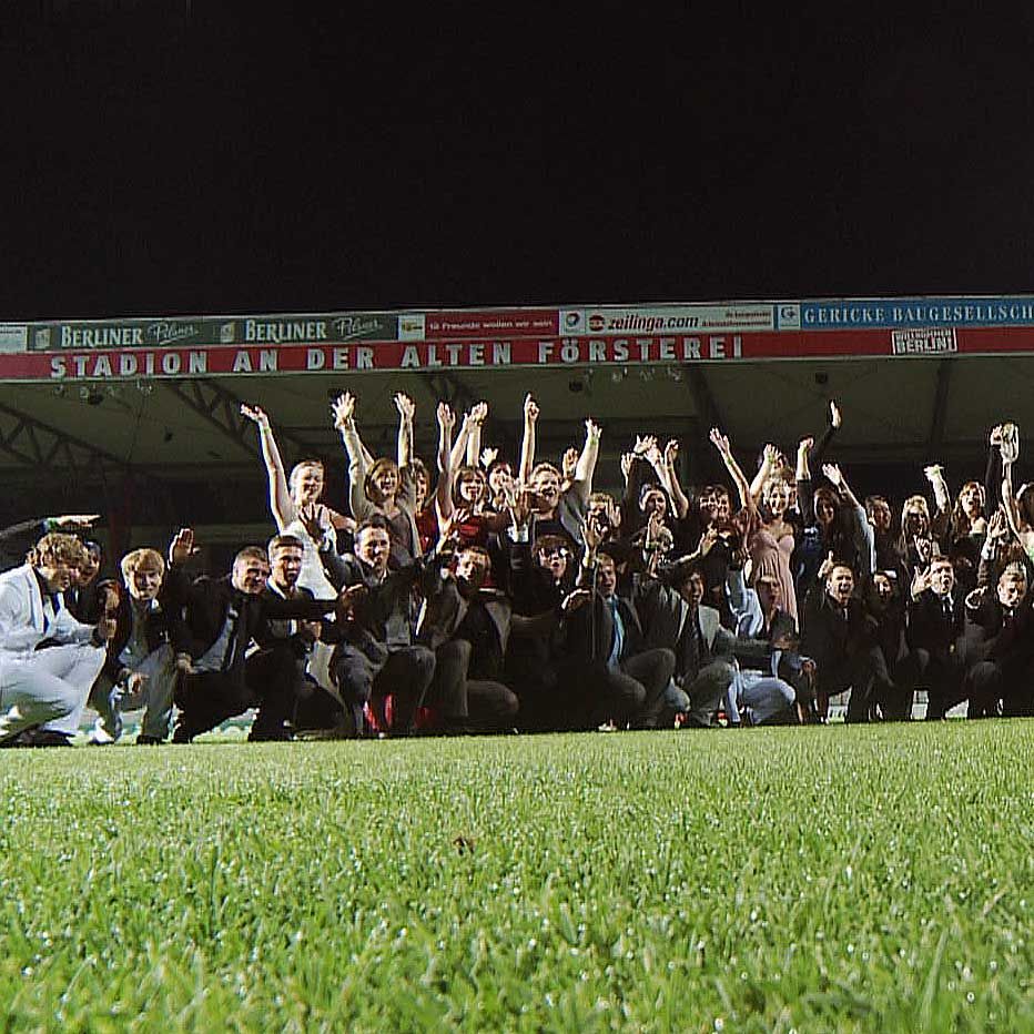 Gruppieren von Menschen auf einem Fußballfeld, die die Arme heben und zur Kamera lächeln, bei Nacht.