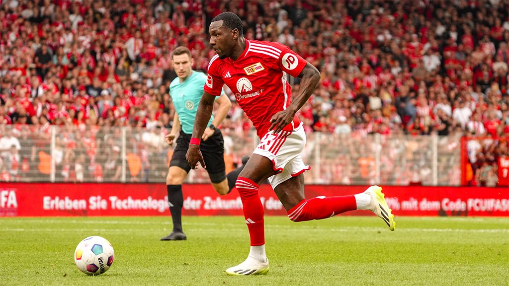 A football player in a red jersey runs with the ball while a referee observes in the background. Fans are visible in the stadium.