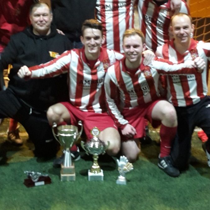 Männer in Fußballtrikots posieren mit Pokalen auf einem Rasenfeld, lächelnd und stolz.