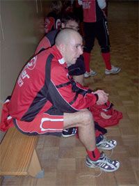 Eine Gruppe von Sportlern in roten Outfits sitzt an der Wand in einer Sporthalle, während sie sich vorbereiten.