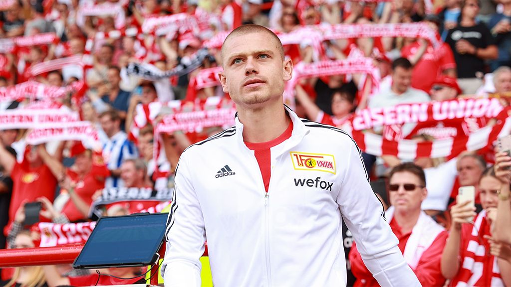 Ein Spieler in einem weißen Trainingsanzug steht in einem Stadion, umgeben von jubelnden Fans mit roten Schals.