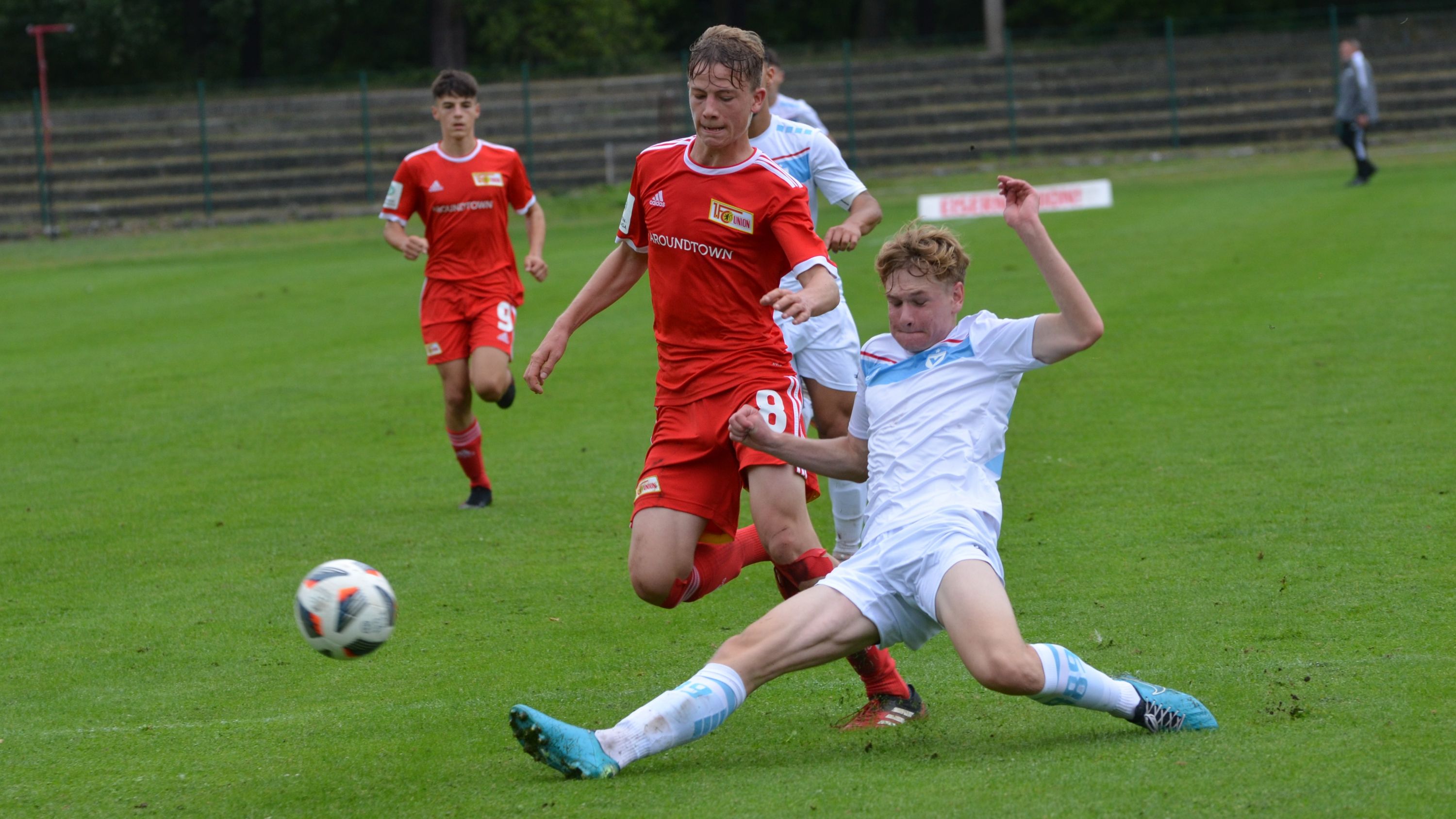 Zwei Fußballspieler kämpfen um den Ball auf einem Rasenfeld, während ein dritter Spieler im Hintergrund läuft.