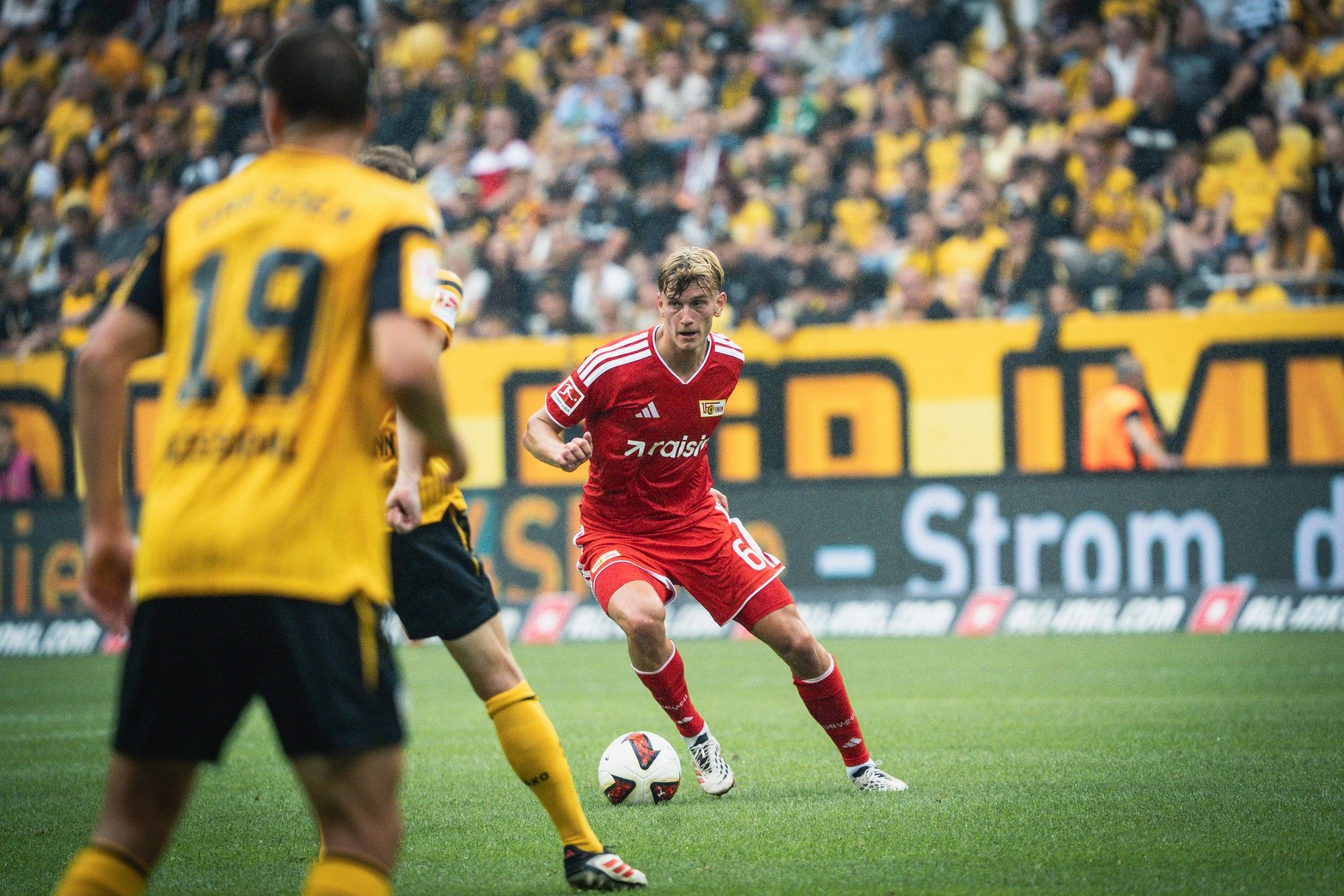 Ein Fußballspieler in roter Ausrüstung dribbelt den Ball, während ein Gegner in gelbem Trikot nah bei ihm steht. Stadionatmosphäre im Hintergrund.