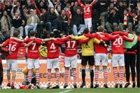 Fußballmannschaft in roten Trikots, die sich nach einem Spiel feiernd umarmen, Fans jubeln im Hintergrund.