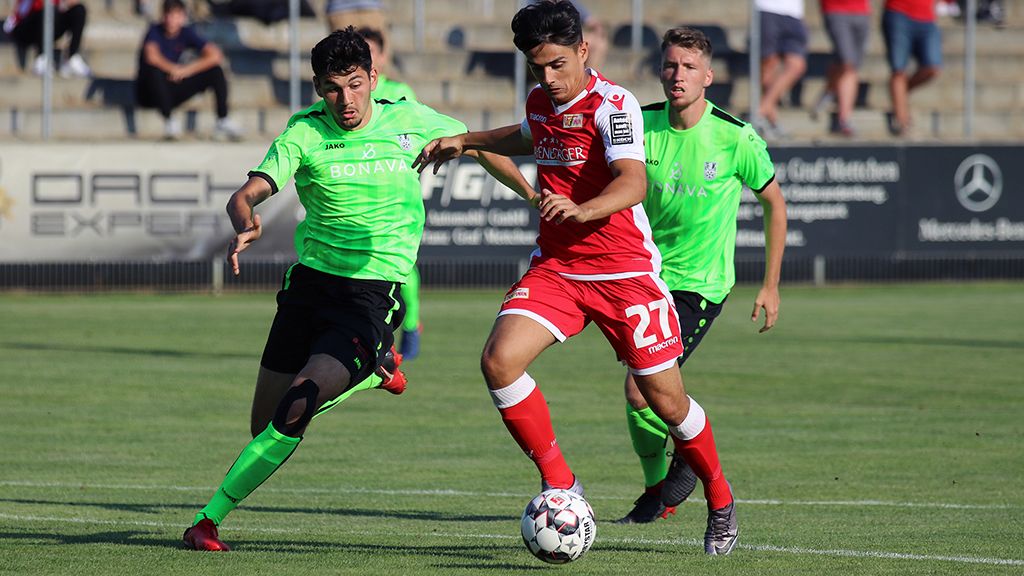Zwei Fußballspieler in grünen Trikots greifen einen Spieler in rotem Trikot an, während dieser den Ball am Spielfeldrand kontrolliert.