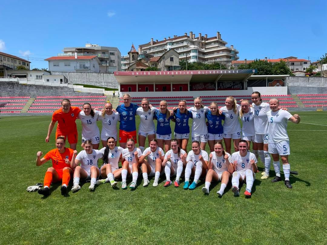 Frauenfußballmannschaft posiert auf dem Rasen, umgeben von einem Stadion. Sunniges Wetter und sportliche Kleidung sind sichtbar.