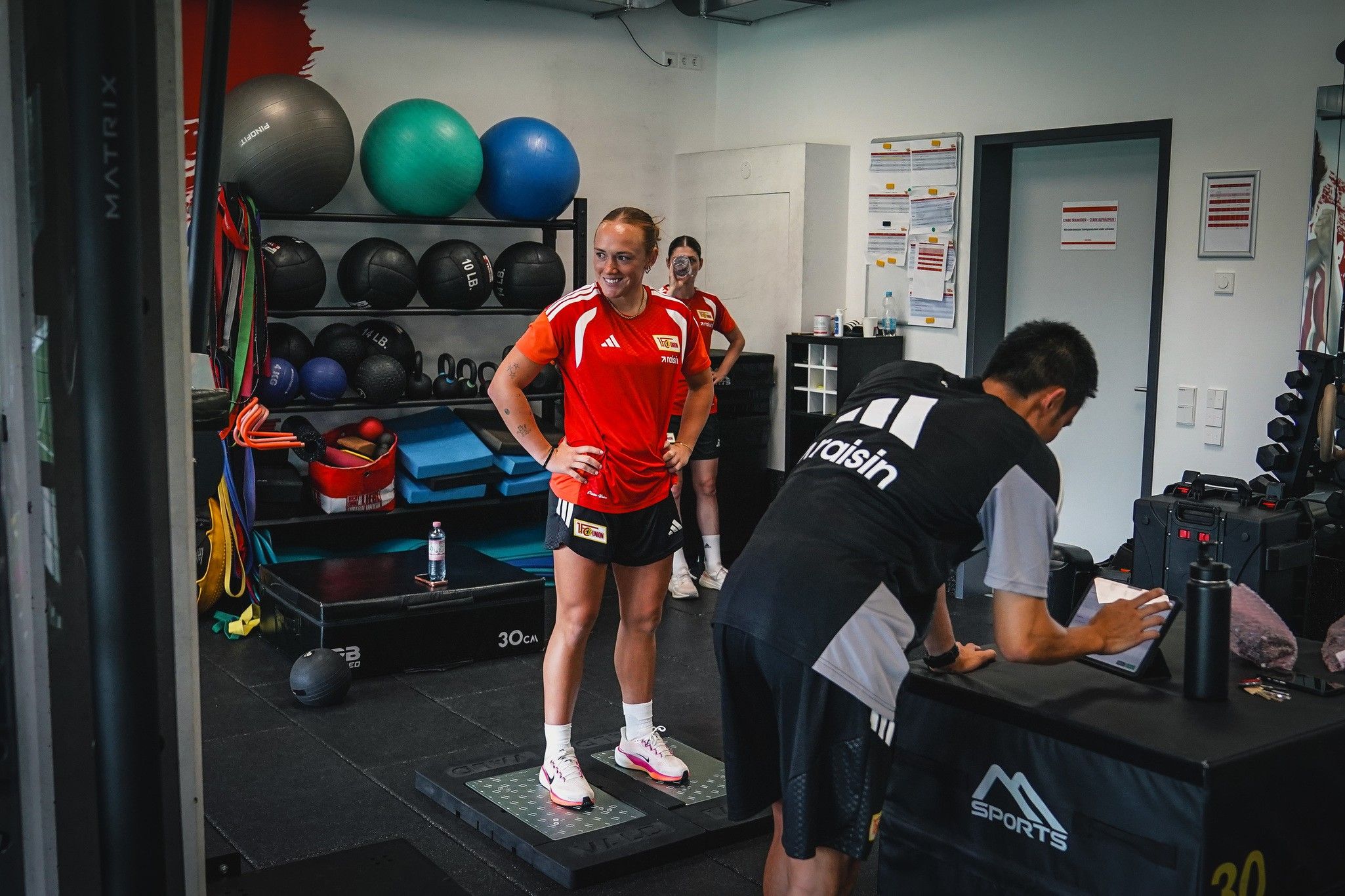 Sportler in Trainingskleidung steht auf einer Waage, während ein Trainer Notizen macht. Fitnessgeräte im Hintergrund.