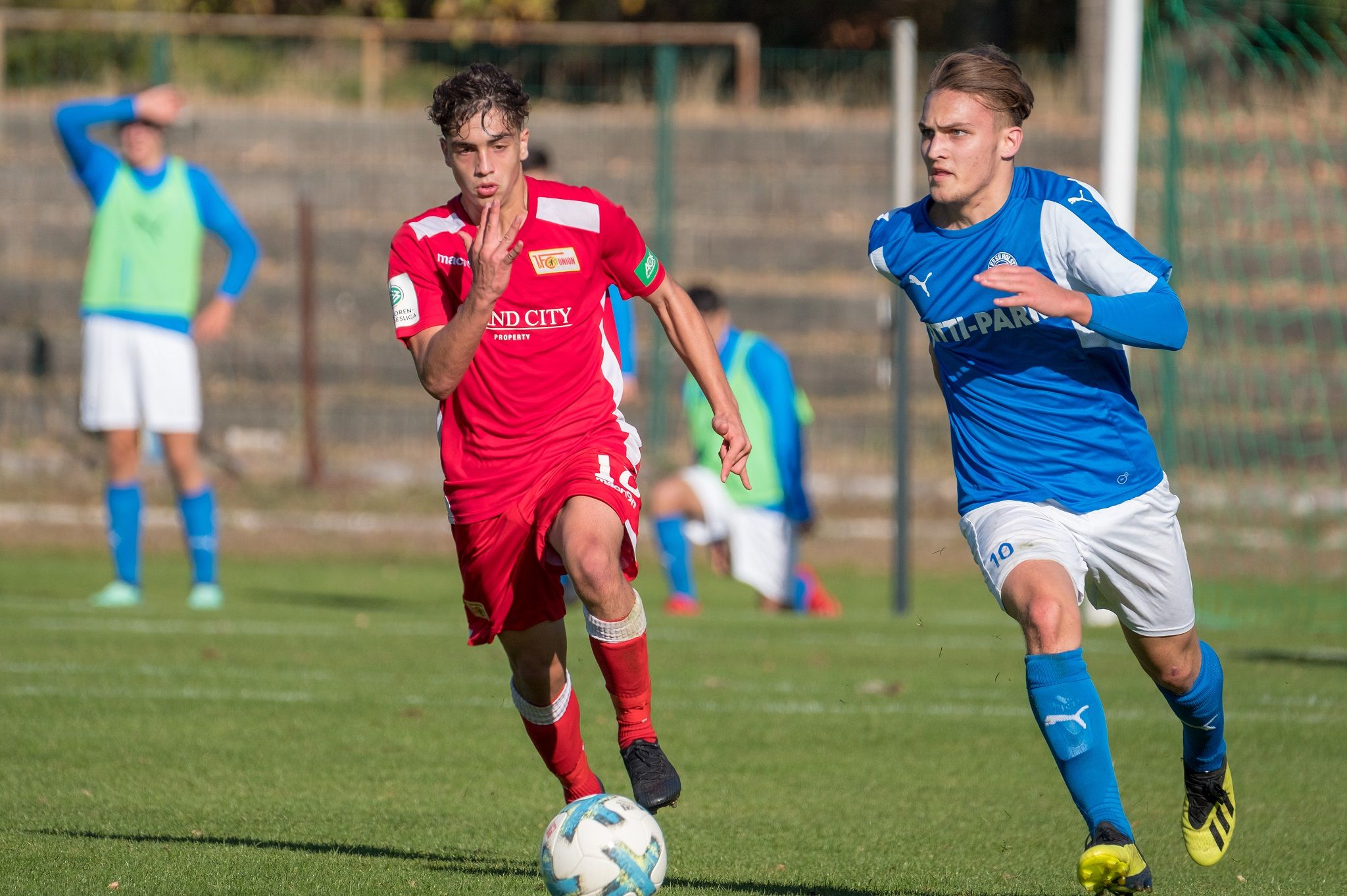 Zwei Fußballspieler in Aktion: Ein Spieler in roter Uniform dribbelt den Ball, während ein Gegner in blauer Uniform folgt.