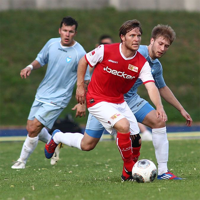 Ein Fußballspieler in rotem Trikot dribbelt den Ball, während zwei Verteidiger in blauen Trikots versuchen, ihn zu stoppen.