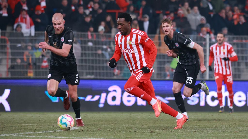 Ein Fußballspieler in rot-weißer Uniform sprintet in Richtung Tor, während zwei Verteidiger im Hintergrund folgen.