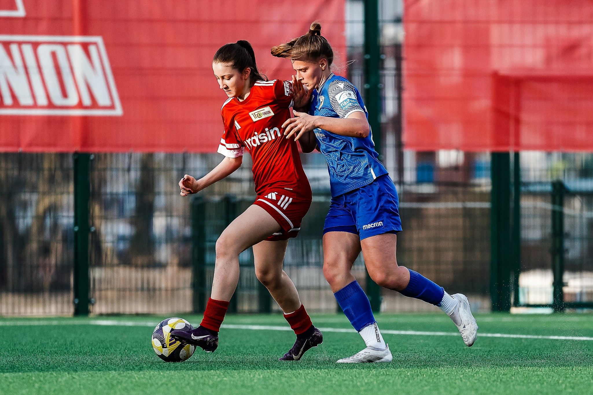 Zwei Frauen im Fußballtrikot spielen an einem Kunstrasenplatz. Eine Spielerin dribbelt den Ball, während die andere sie verfolgt.