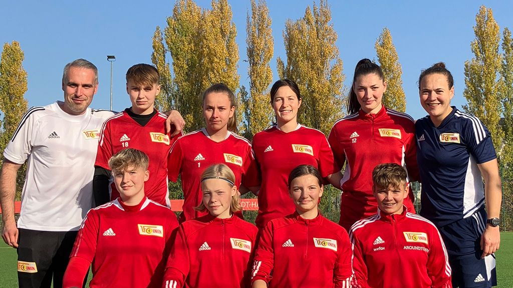 Eine Gruppe von sieben Personen in roten Fußballtrikots, posierend auf einem Platz, umgeben von bunten Bäumen.