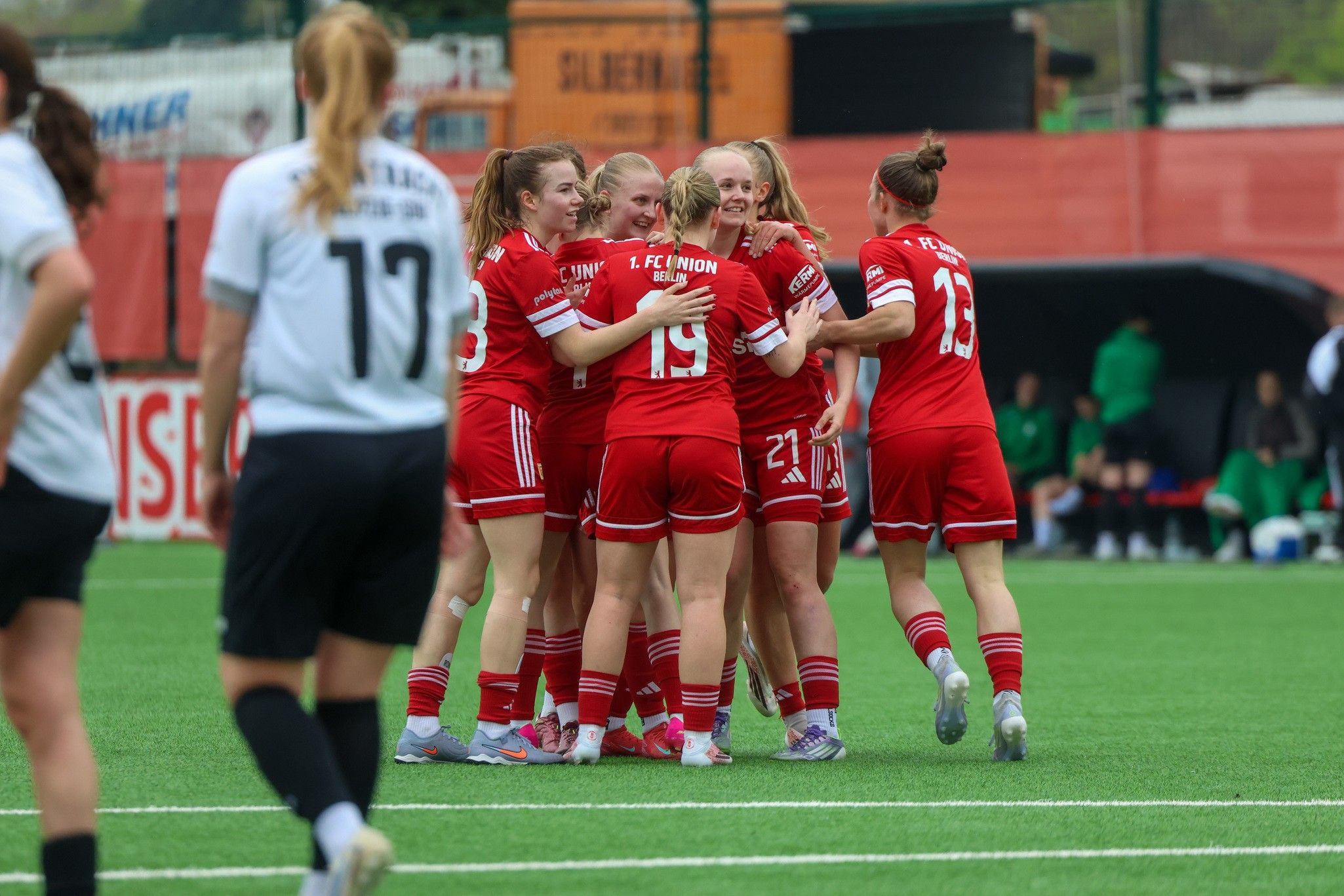 Sechs Spielerinnen in roten Trikots umarmen sich jubilierend auf einem Sportplatz.