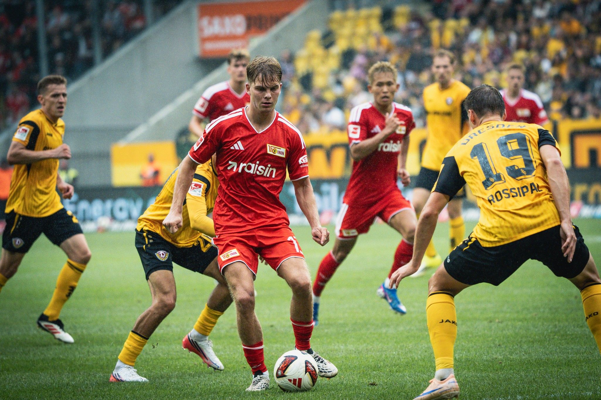 Ein Fußballspieler in rotem Trikot dribbelt den Ball, umgeben von Spielern in gelben und schwarzen Trikots auf dem Spielfeld.