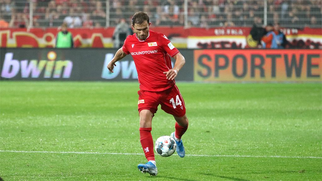 A soccer player in a red uniform with the number 14 dribbles the ball on the field. Background with spectators.