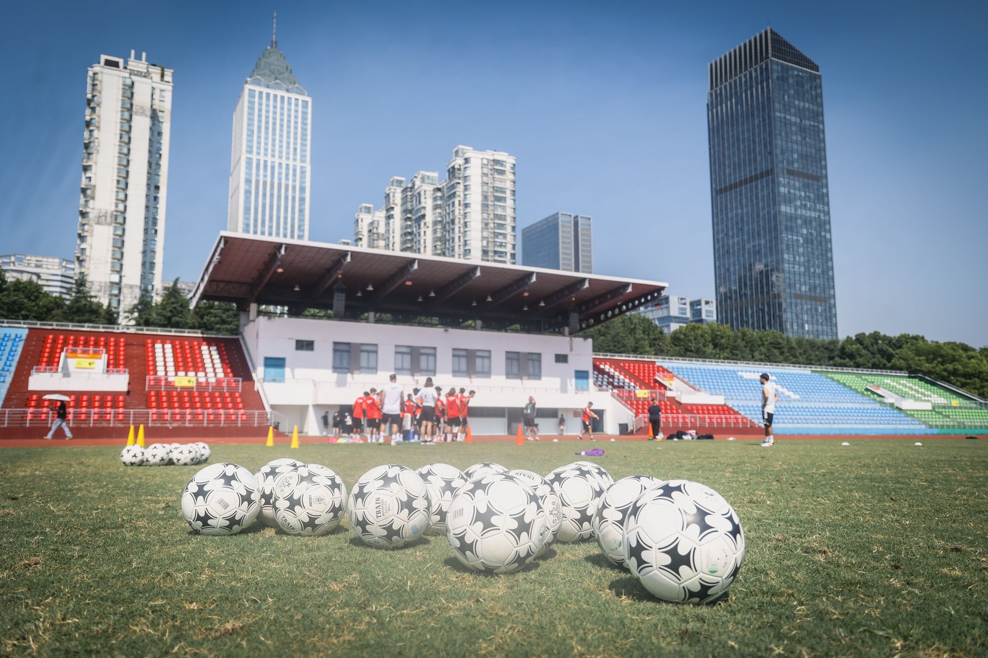 Fussballfeld mit mehreren Bällen im Vordergrund und Spielern in der Trainingssituation vor Stadien der Stadt.