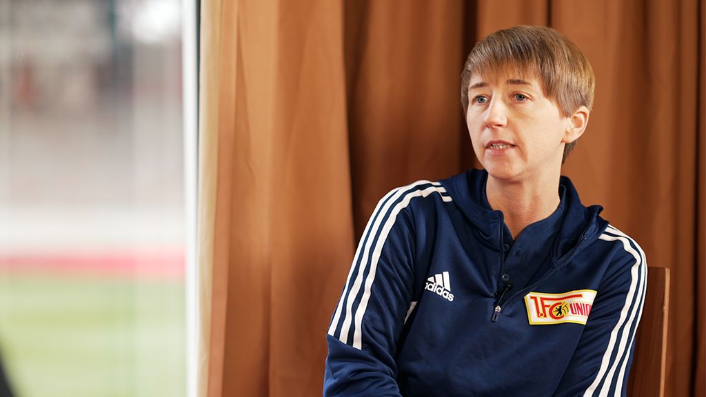 Eine Person mit kurzer Frisur trägt eine Sportjacke und spricht in einem Raum mit braunen Vorhängen.