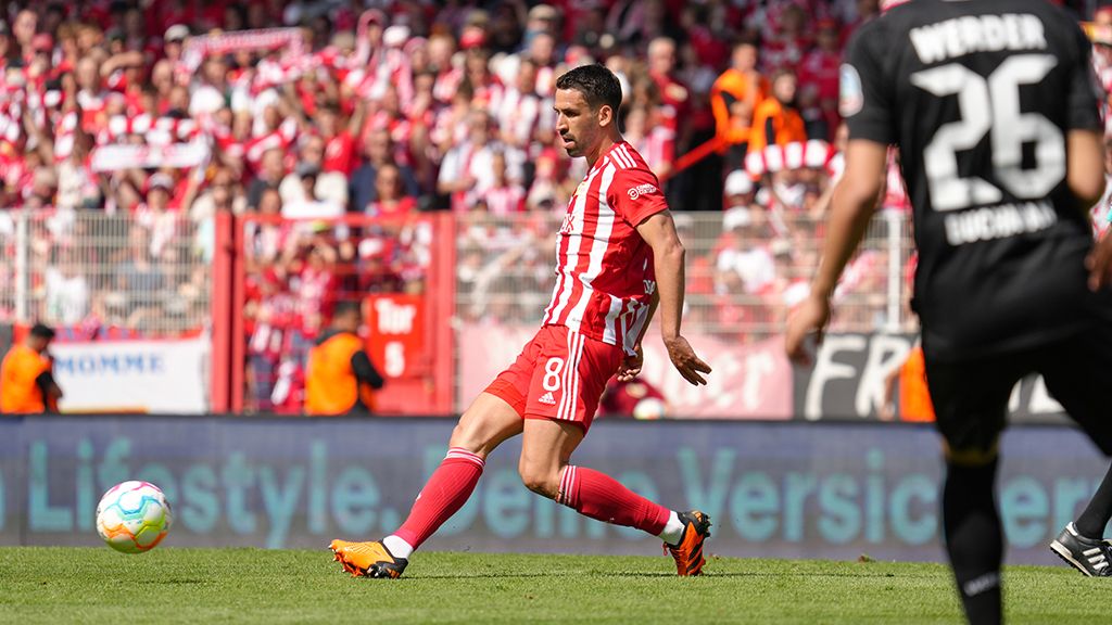 Spielszene im Fußballstadion mit einem Spieler in rotem Trikot, der den Ball schießt, während Fans im Hintergrund jubeln.