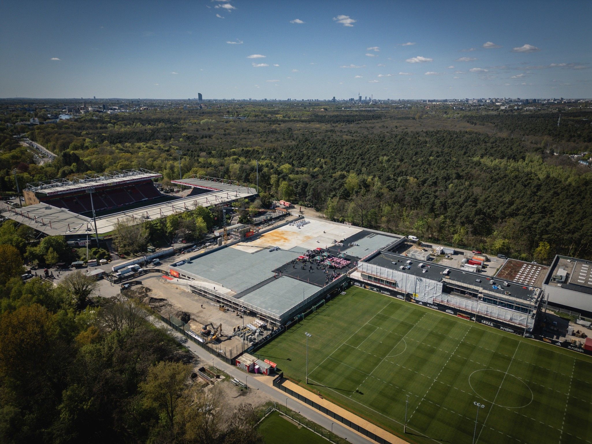 Luftaufnahme eines Sportgeländes mit einem Fußballfeld, einem Stadion links und angrenzendem Waldgebiet im Hintergrund.