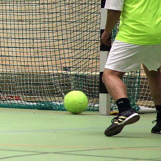 Ein Sportler in hellgrüner Trikotfarbe schießt einen gelben Ball in Richtung des Torraums auf einem Hallenboden.