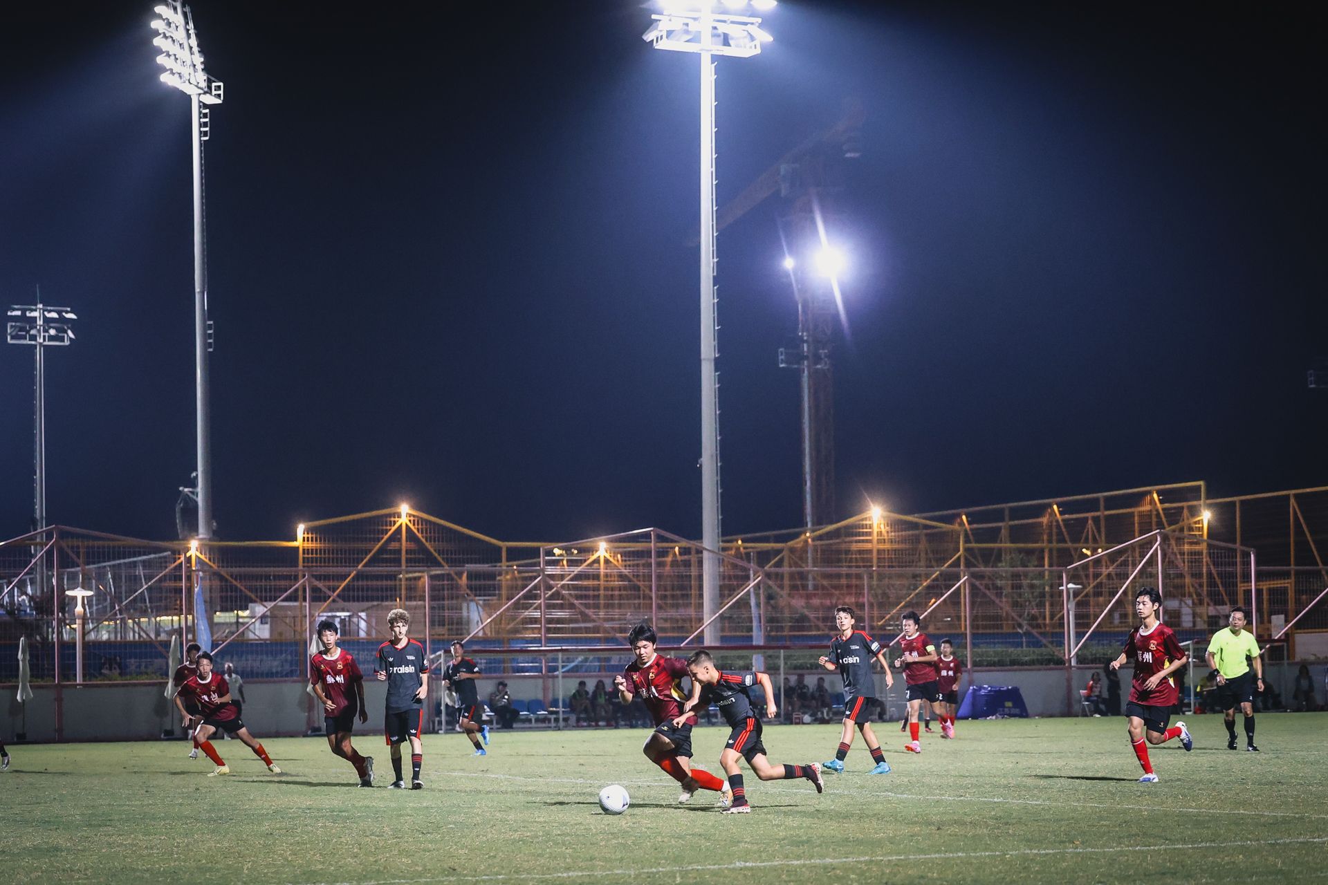 Fußballspiel bei Nacht mit Spielern in roten und schwarzen Trikots, die einem Ball auf einem gut beleuchteten Feld nachjagen.