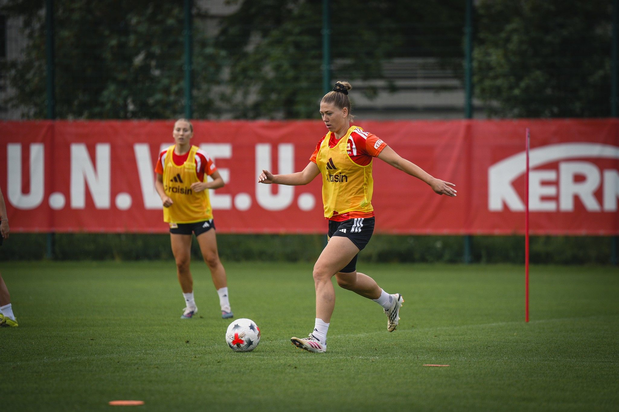 Eine Fußballspielerin schießt den Ball während des Trainings. Im Hintergrund sind weitere Spielerinnen und ein Trainingsfeld zu sehen.