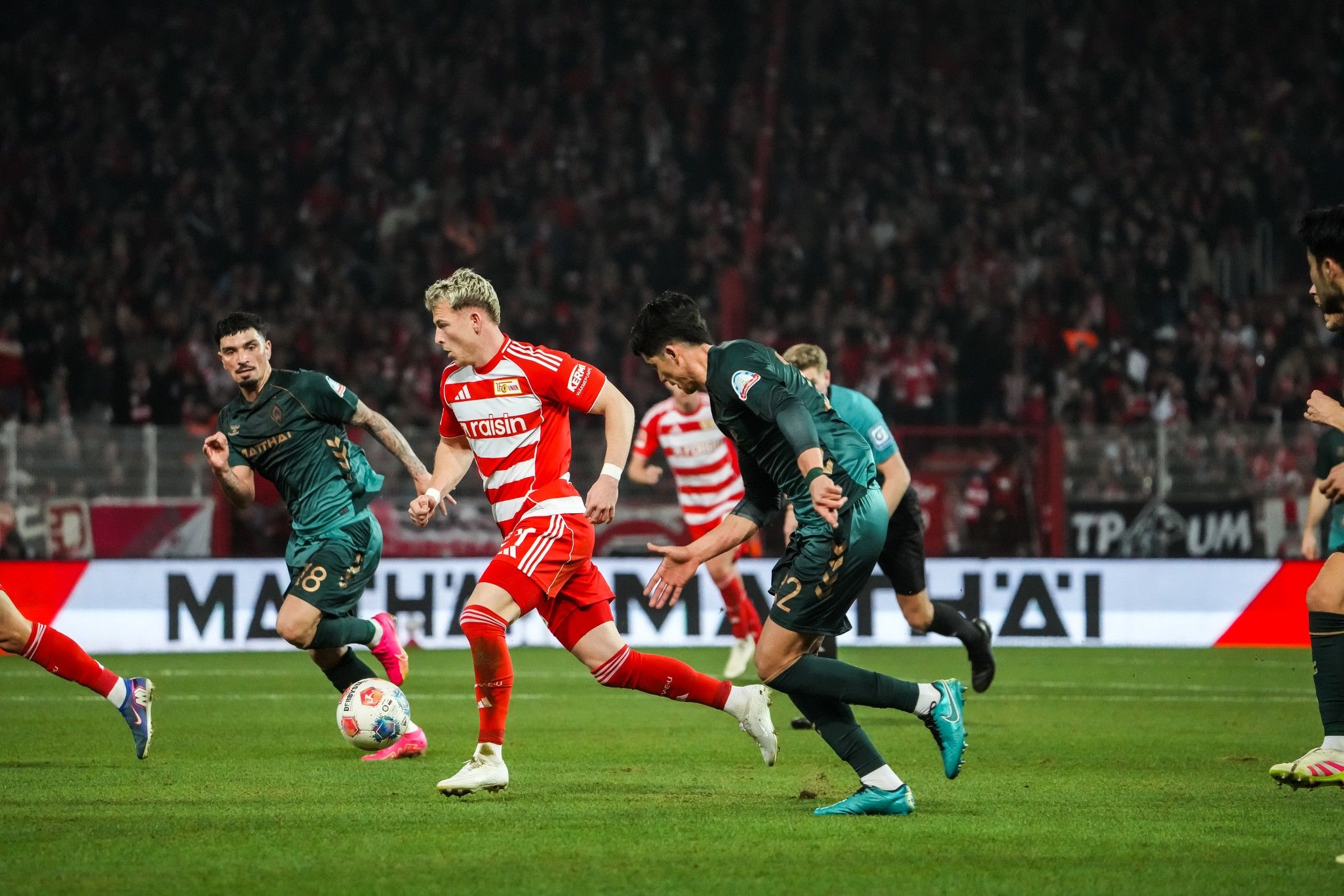 Ein Fußballspiel, bei dem Spieler in roten und grünen Trikots um den Ball kämpfen, vor einer jubelnden Menge.