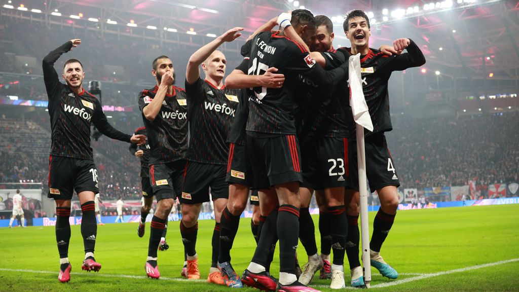 Fußballmannschaft jubelt nach einem Tor, Spieler umarmen sich auf dem Spielfeld. Stadion ist voll mit Fans.
