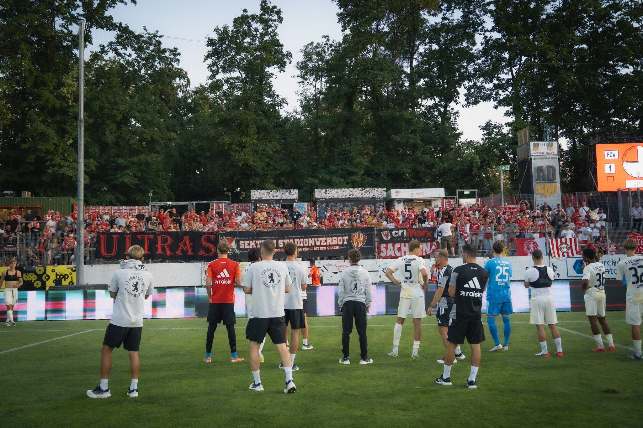 Fußballmannschaft steht auf dem Spielfeld vor einer jubelnden Menge mit transparentem Banner im Hintergrund.
