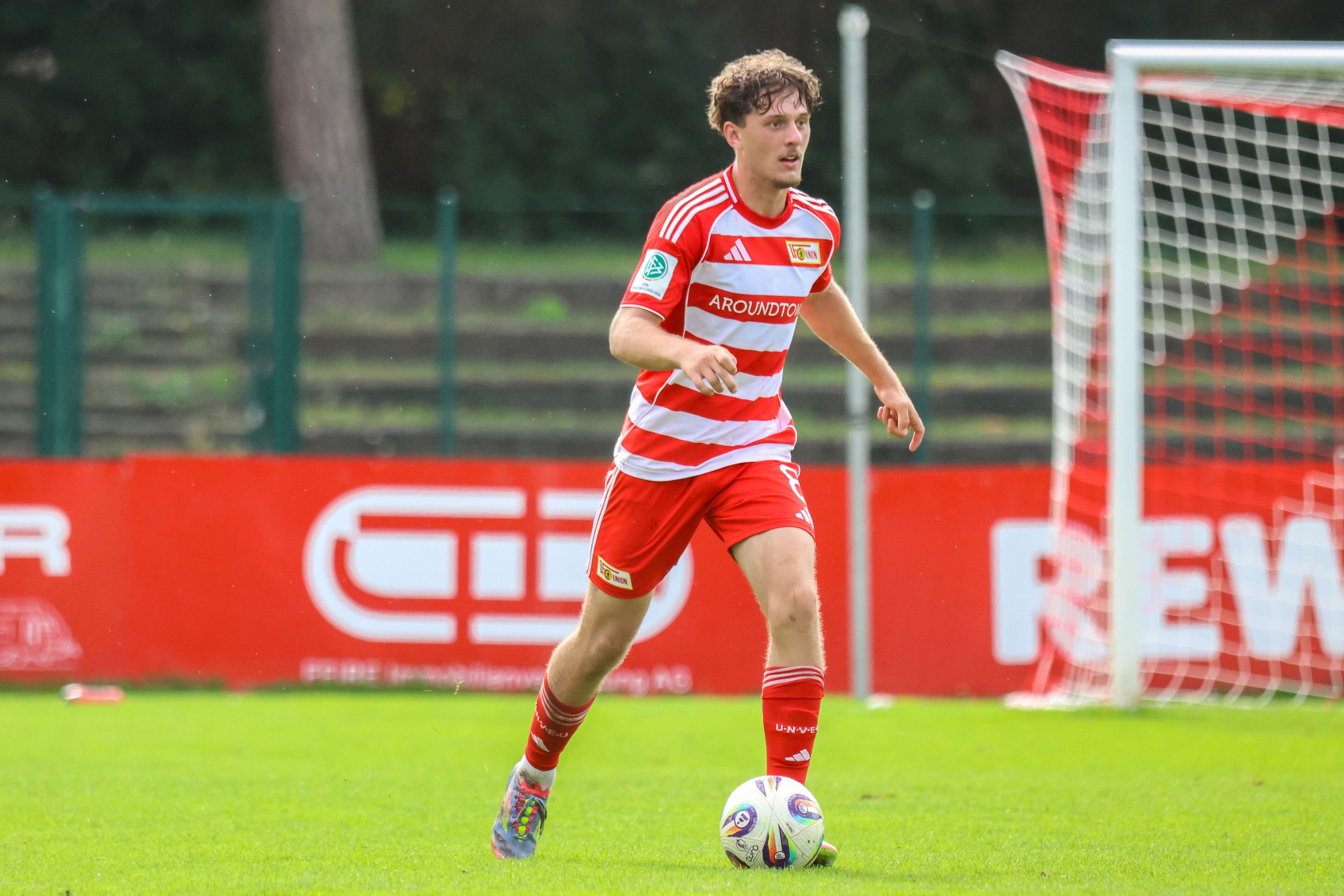 Ein Fußballspieler in rot-weiß gestreifter Kleidung dribbelt den Ball auf einem Trainingsplatz. Im Hintergrund Tore und Werbebanner.