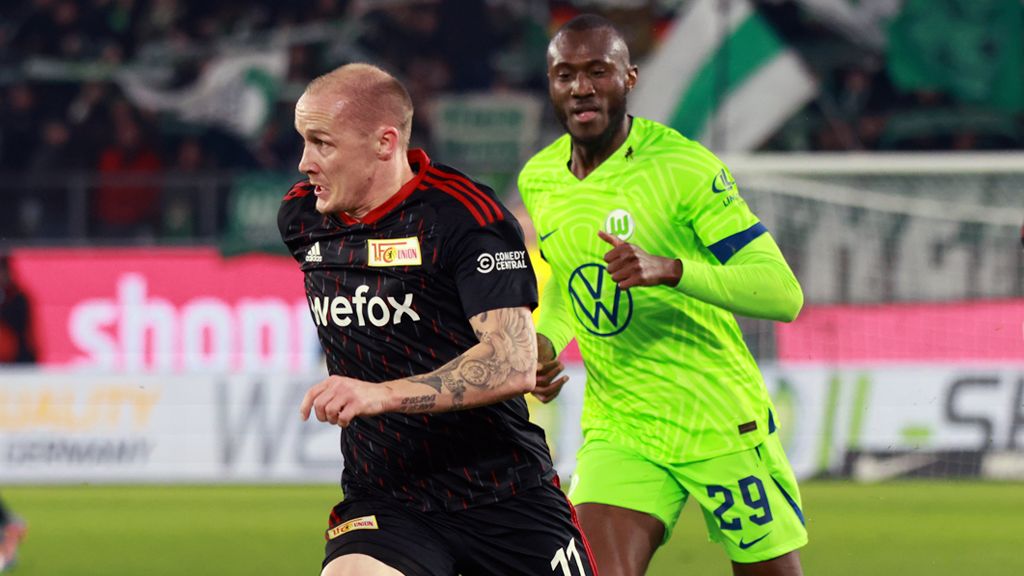 Zwei Fußballspieler in Aktion während eines Spiels, einer in rot-schwarz, der andere in grün mit einer Kapitänsbinde.