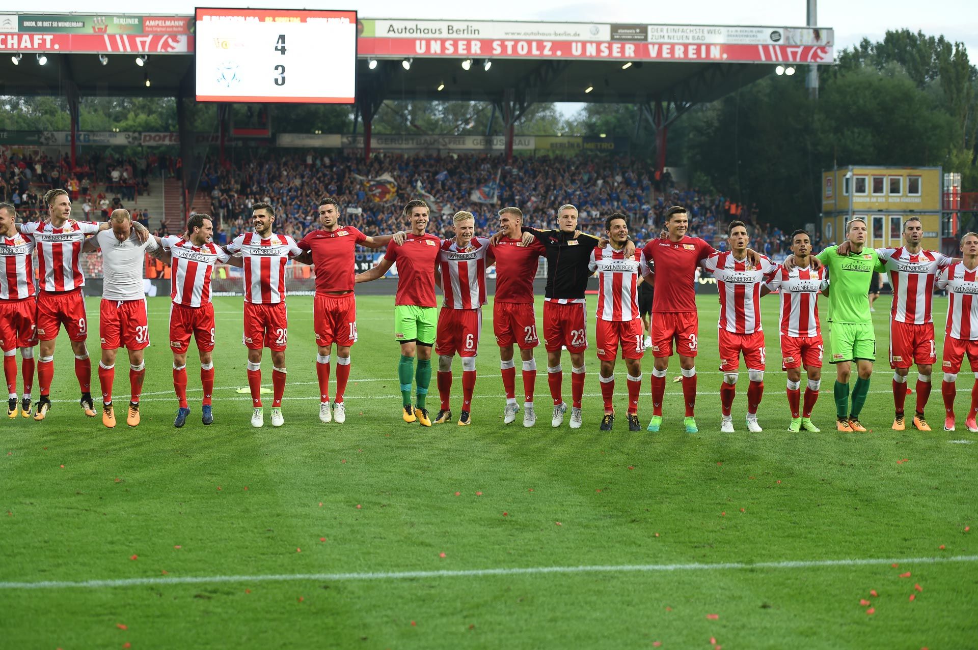 Die Fußballmannschaft steht auf dem Platz. Sie freuen sich über den Sieg. Im Hintergrund jubeln die Fans.
