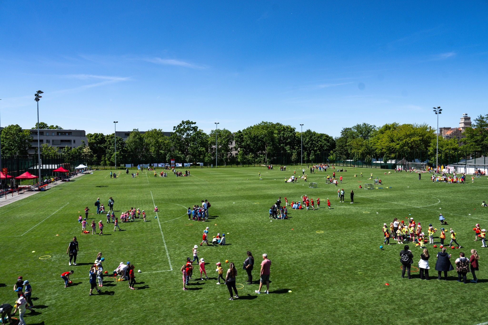 Kindersportveranstaltung auf einem weitläufigen, grünen Rasenplatz mit vielen Jugendlichen und mehreren Spielstationen.