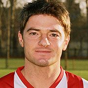 Ein Fußballspieler mit kurzen, dunklen Haaren, trägt ein rotes und weißes Trikot, steht vor einem unscharfen grünen Hintergrund.