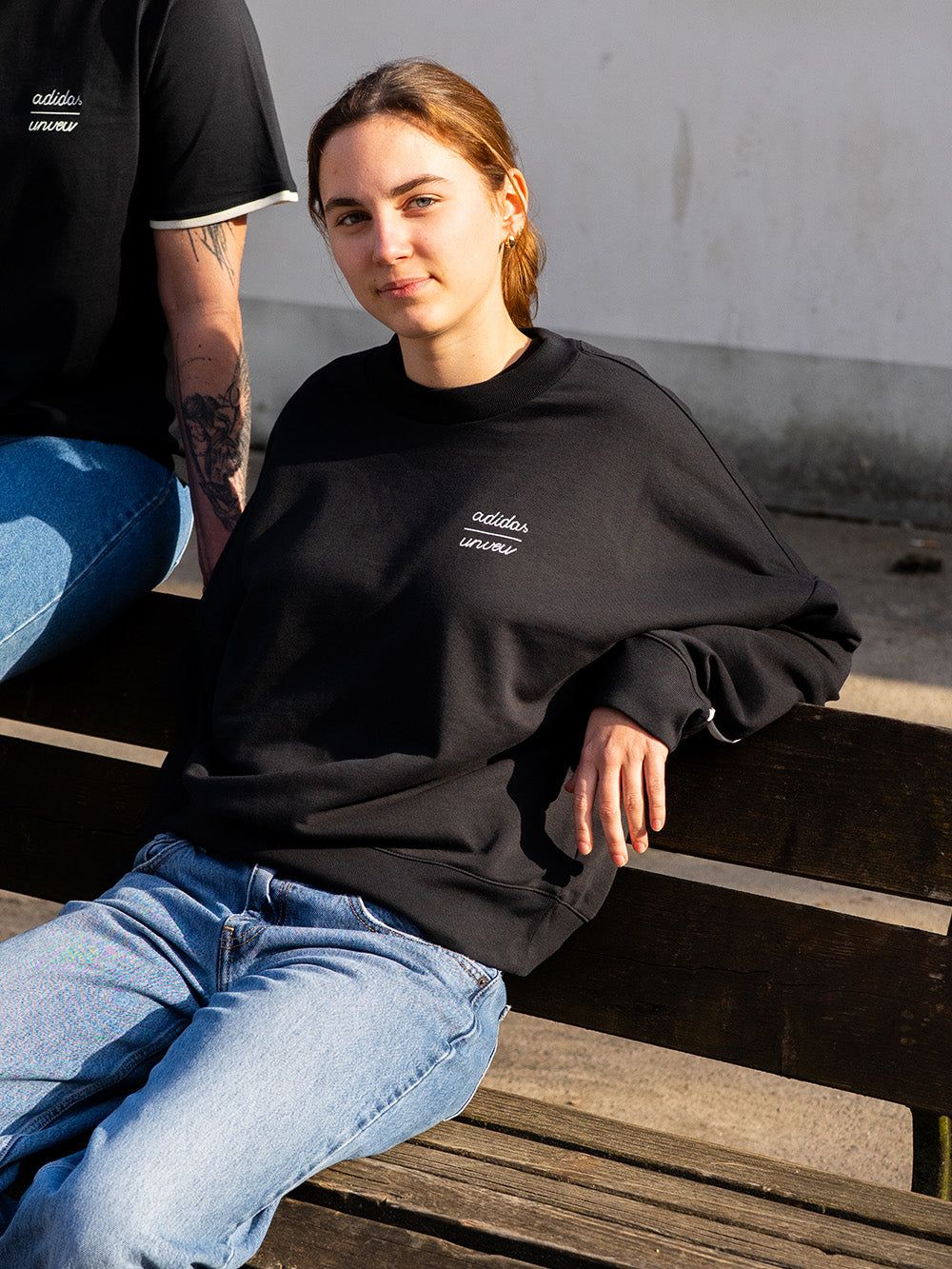 Eine junge Frau sitzt auf einer Bank, trägt einen schwarzen Sweatshirt und Jeans, mit einem neutralen Hintergrund.