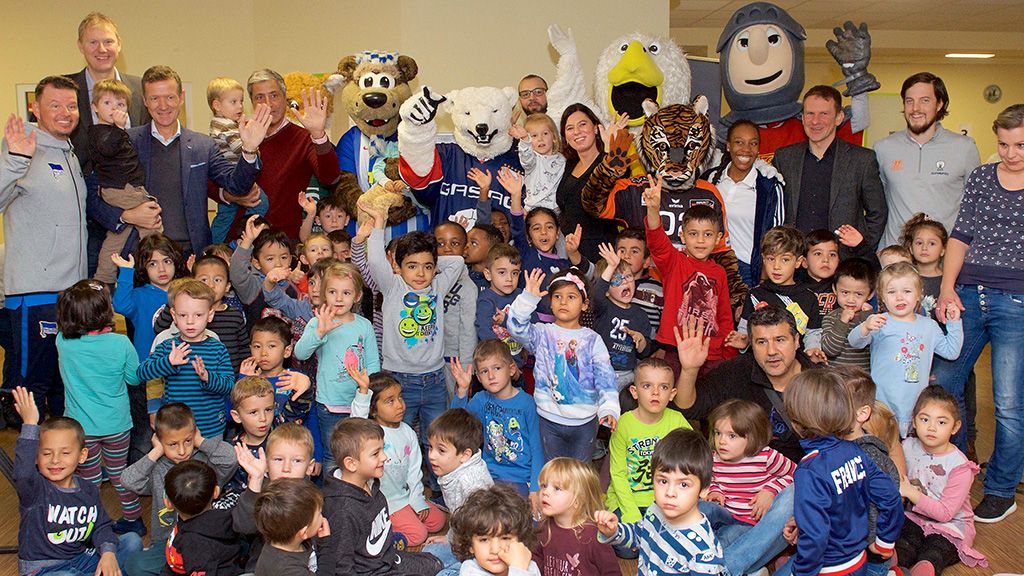 Eine große Gruppe von Kindern und Erwachsenen, darunter Maskottchen, posiert lächelnd für ein Foto in einem Raum.