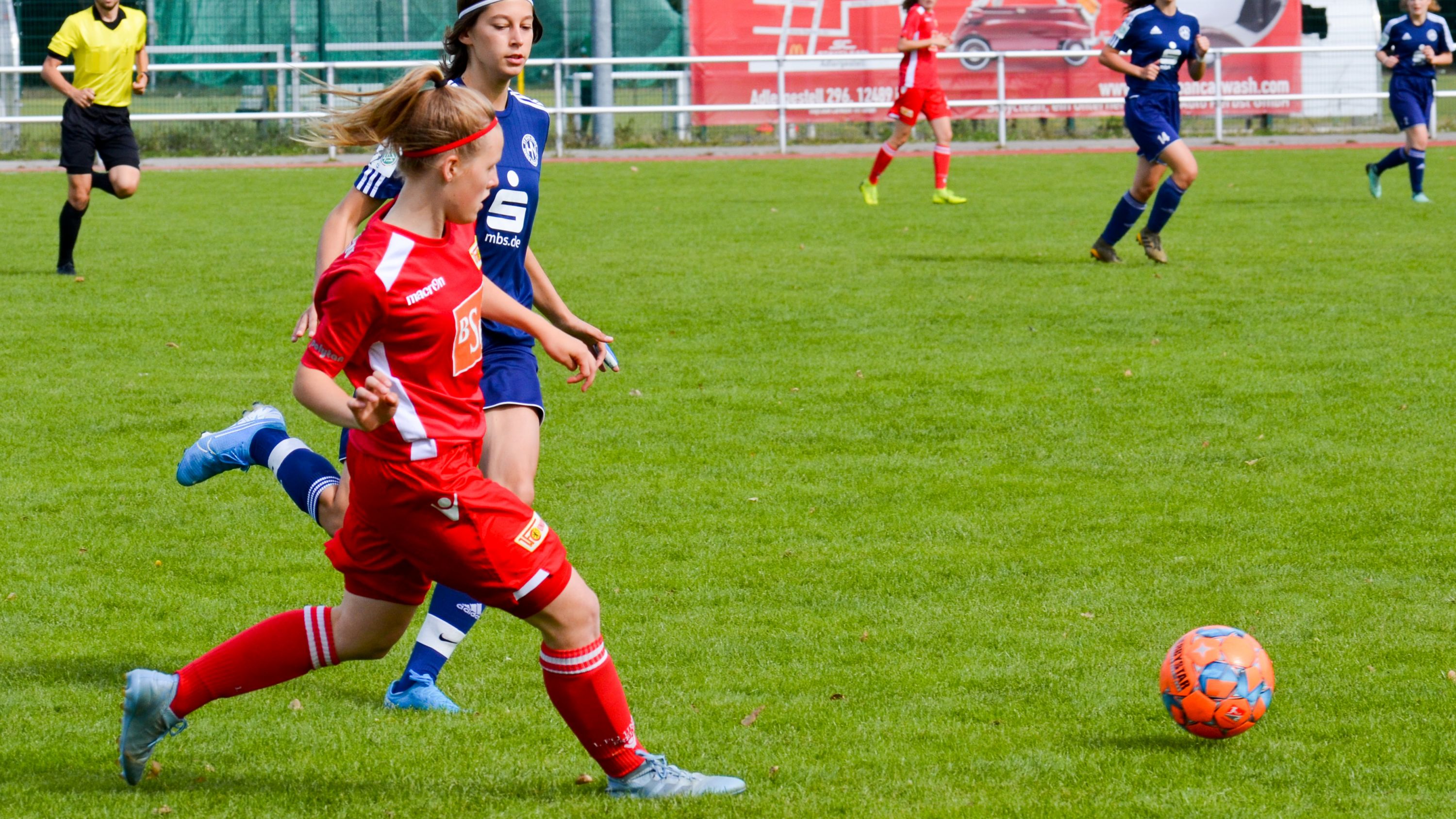 Eine Spielerin in rotem Trikot kickt den Ball, während eine Gegnerin in blauem Trikot nahe dabei ist. Sportplatz im Hintergrund.