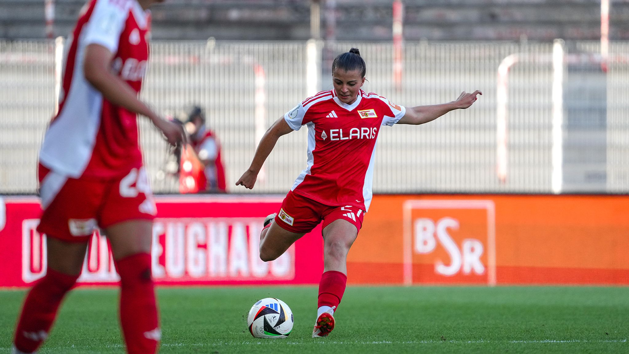 Spielerin in rotem Trikot schießt einen Fußball. Im Hintergrund ist ein weiteres Teammitglied sichtbar.