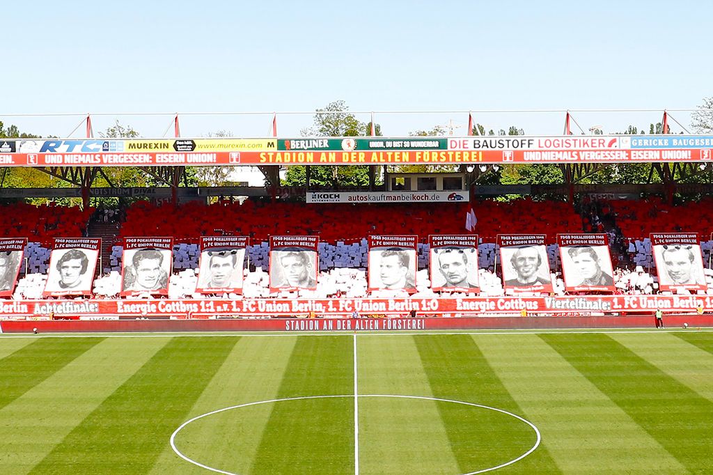 Ein Fußballstadion mit roten und weißen Fanschals, großformatigen Porträts von Spielern und einem grünen Spielfeld.