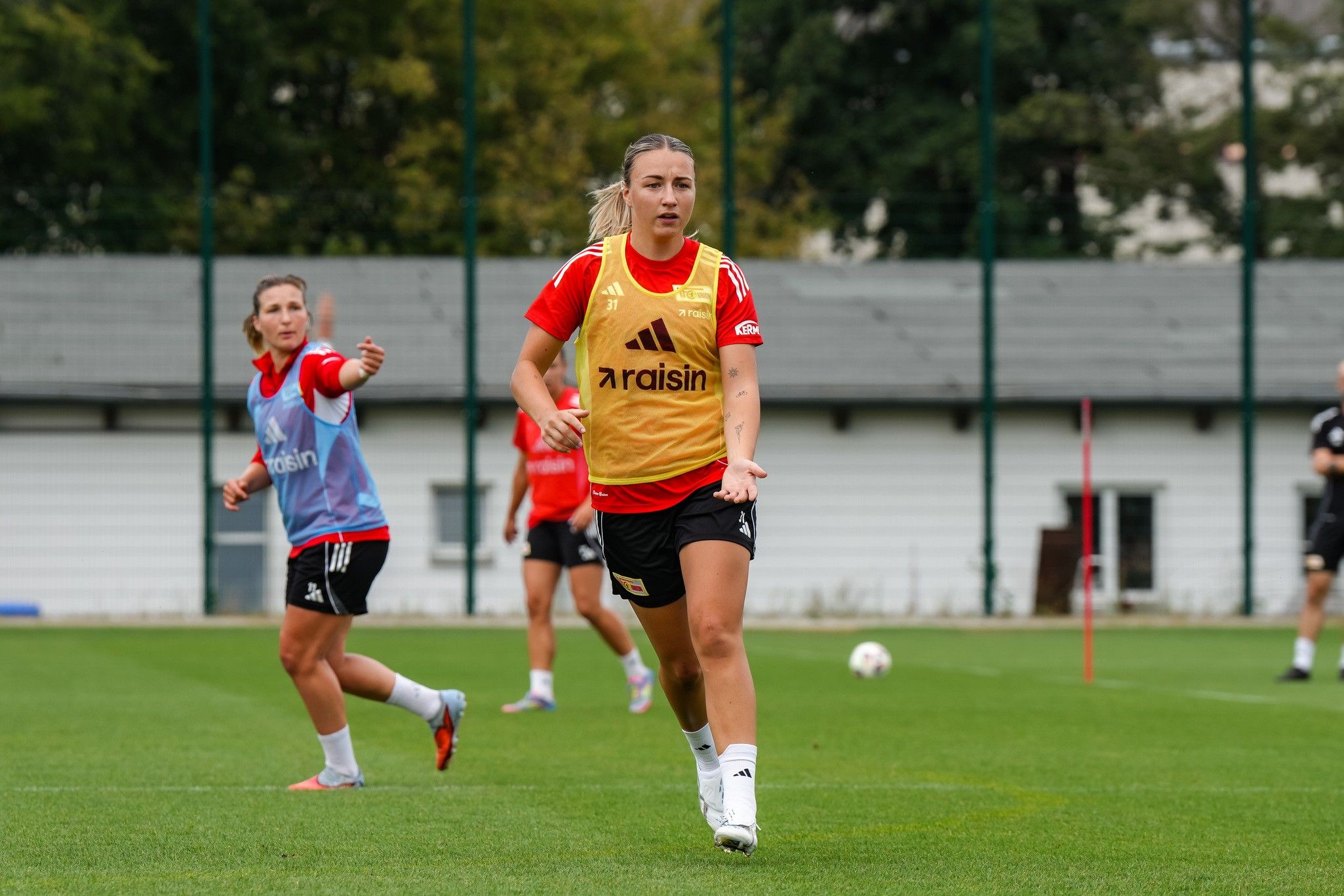 Frauenfußballspielerinnen trainieren auf einem Rasenplatz, eine in gelbem Trikot läuft, die andere gibt Anweisungen.