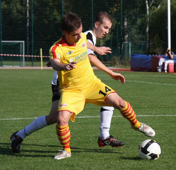 Ein Fußballspieler in gelber Uniform mit rotem Streifen tritt den Ball, während ein weiterer Spieler ihn von hinten verfolgt.