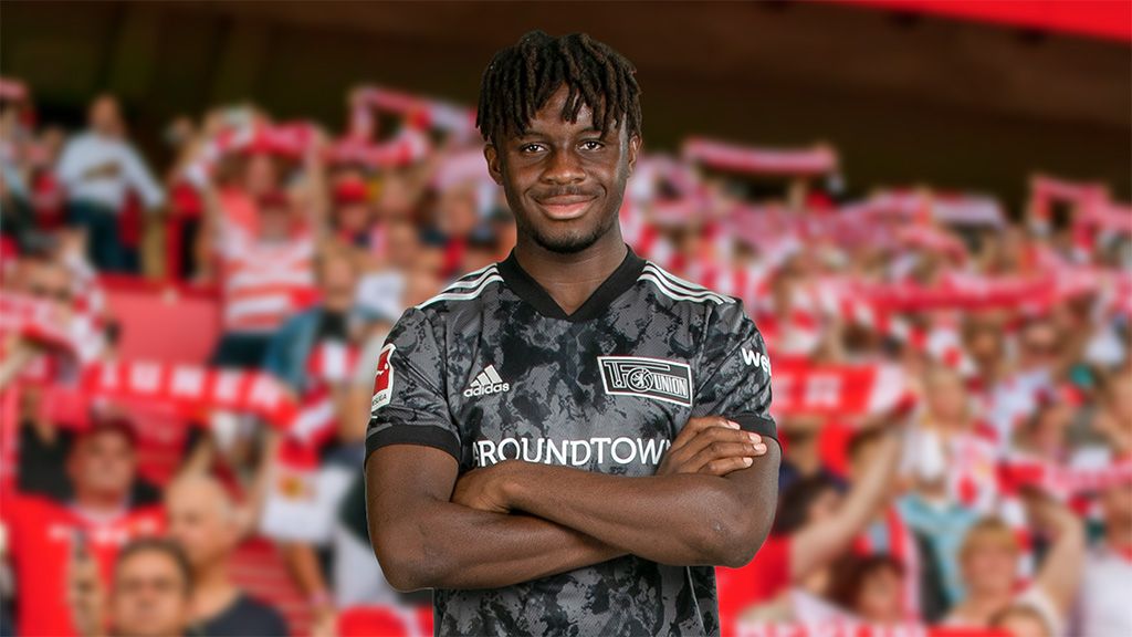 Ein Fußballspieler in einem grauen Trikot steht mit verschränkten Armen vor einer jubelnden Menge im Stadion.