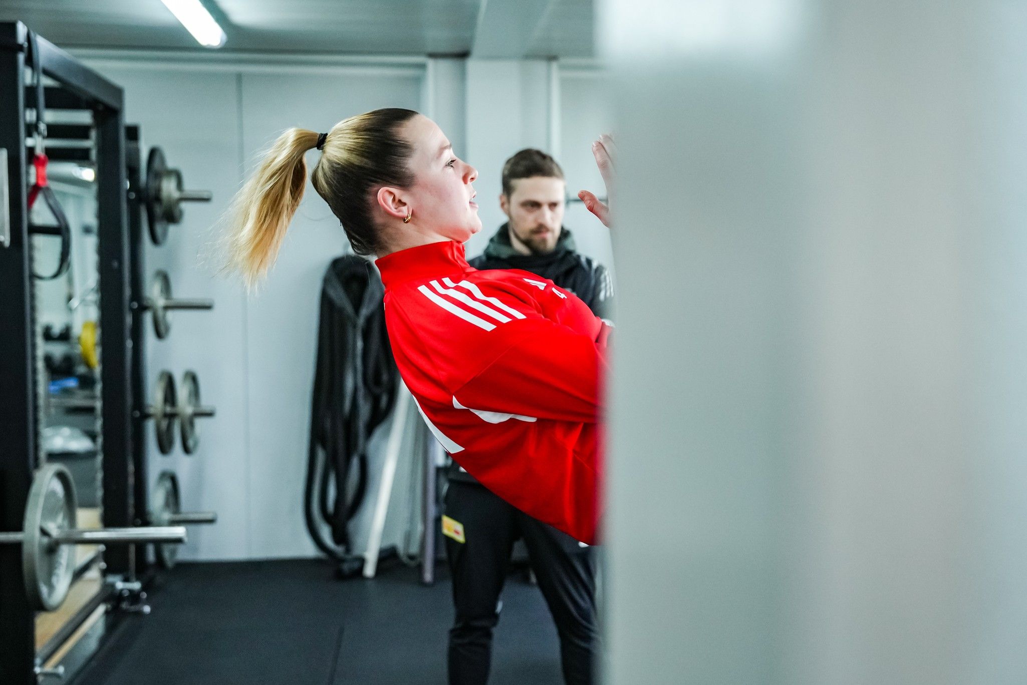 Eine Person in einem roten Trainingsanzug führt eine Übung im Fitnessstudio durch, während eine zweite Person im Hintergrund zuschaut.