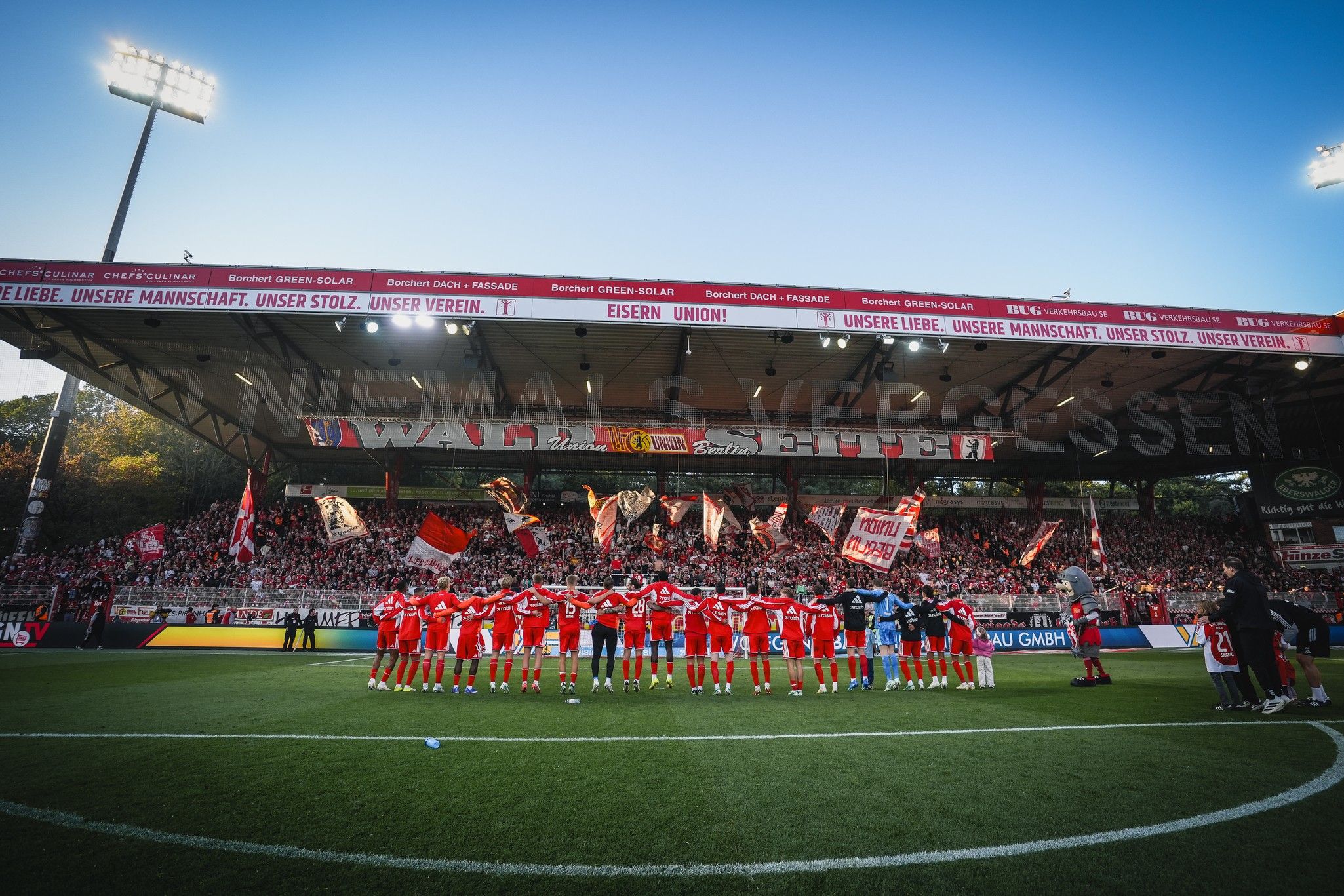 Eine Fußballmannschaft steht auf dem Platz und feiert, während Fans im Hintergrund Transparente und Flaggen halten.