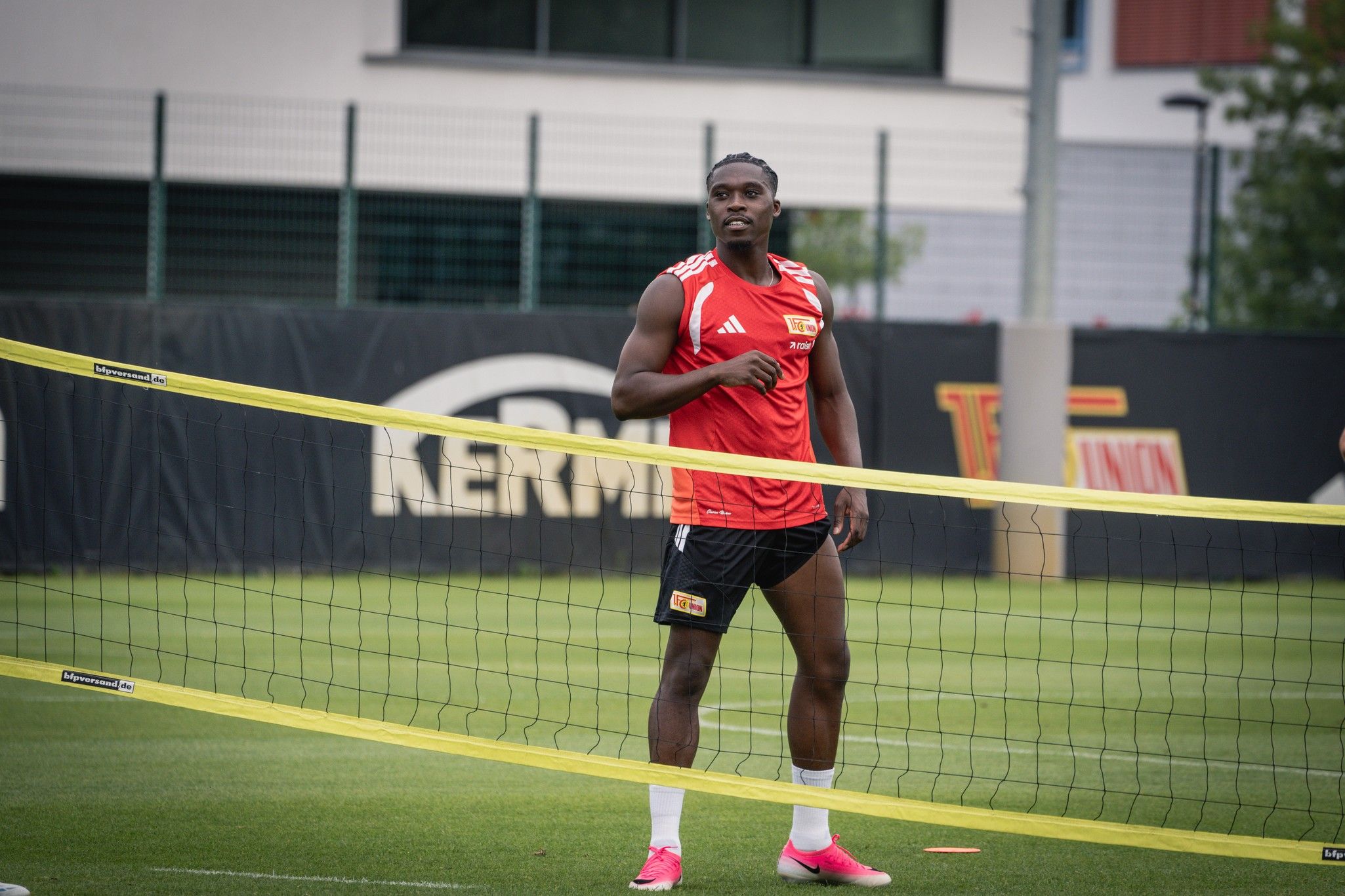 Ein Fußballspieler steht neben einem Tennisnetz auf einem Trainingsplatz. Er trägt ein rotes Trikot und schwarze Shorts.