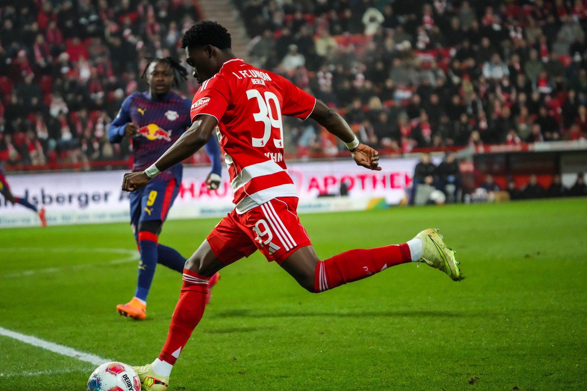 Ein Fußballspieler in rot-weißer Kleidung dribbelt den Ball, während ein Gegner in dunklerer Kleidung ihn beobachtet. Stadion im Hintergrund.