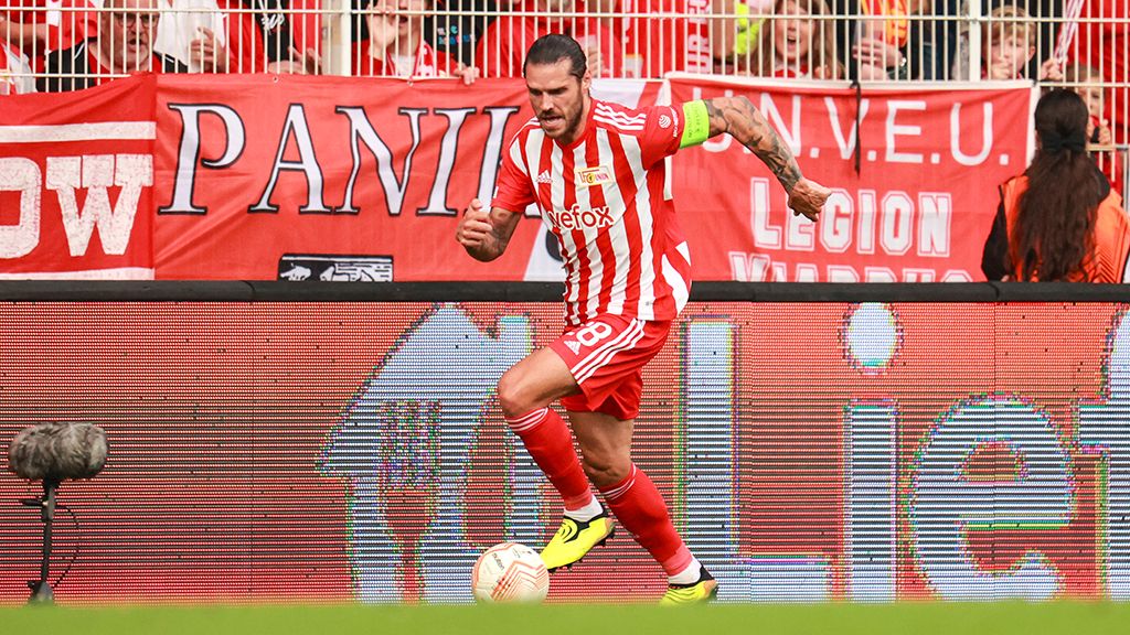 Ein Spieler in rot-weißer Fußballausrüstung dribbelt den Ball im Stadion, umgeben von jubelnden Fans und Vereinsfahnen.