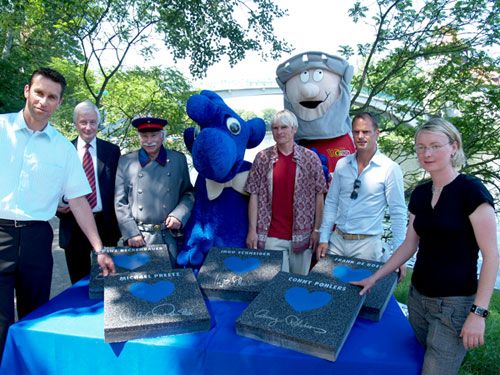 Eine Gruppe von Menschen sowie ein blauer, cartoonartiger Charakter posieren vor Gedenksteinen in einem Freiluftumfeld.